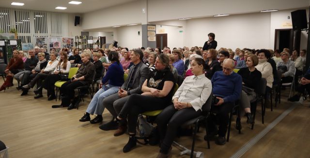 Na zdjęciu znajduje się publiczność na krzesłąch na holu biblioteki. W tle znajduje się stojąca kobieta.