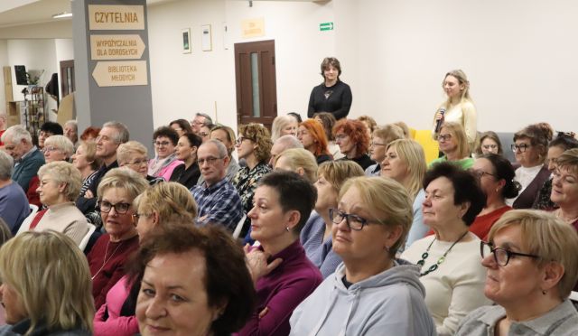 Na zdjęciu znajduje się widownia na holu biblioteki, w tle znajdują się dwie stojące kobiety, jedna z mikrofonem.