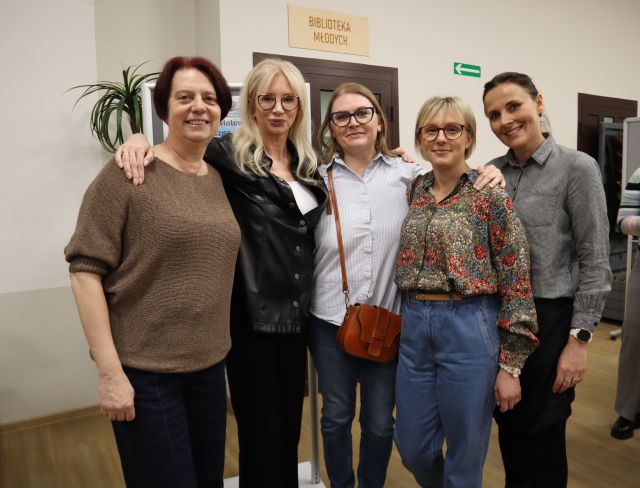 Na zdjęciu znajduje się pięć kobiet stojących wspólnie do zdjęcia. Za kobietami znajdują się drzwi, kawałek zielonej rośliny.