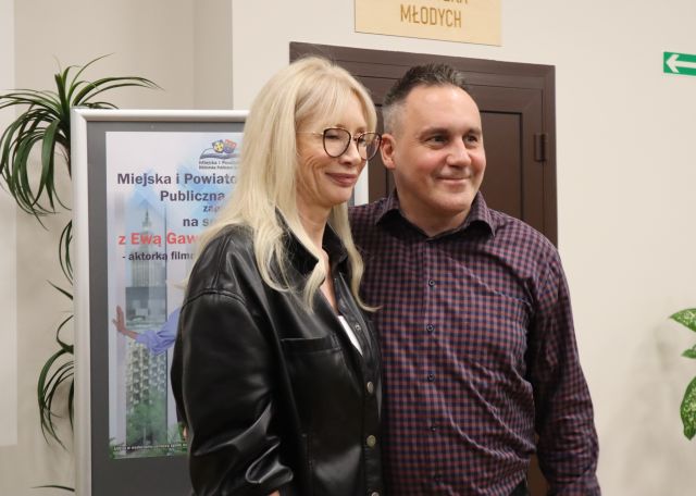 Na zdjęciu znajduje się mężczyzna stojący do zdjęcia wraz kobietą, za nimi znajduje się plakat oraz roślina.