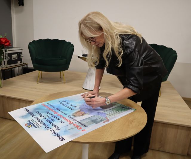 Na zdjęciu znajduje się kobieta nachylająca się przy stoliku aby podpisać plakat promujący spotkanie, za nią znajduje się mała scena z fotelami.
