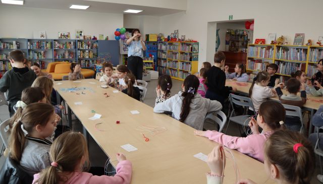 Na zdjęciu w centrum znajdują się połączone stoły przy których siedzą dzieci, obok w tle znajduje się jeszcze jeden ciąg złączonych stolików z resztą uczestników. Pomiędzy stolikami stoi kobieta i pokazuje im jak wykonać breloczek z żyłki. W tle znajdują się półki z książkami.