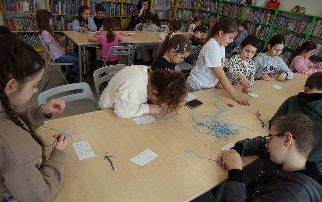 Na zdjęciu znajdują się dzieci przy złączonych stołach, które wykonują breloczki z żyłki, w tle znajduje się jeszcze jeden rząd złączonych stolików z uczestnikami, a za nimi półki z książkami.