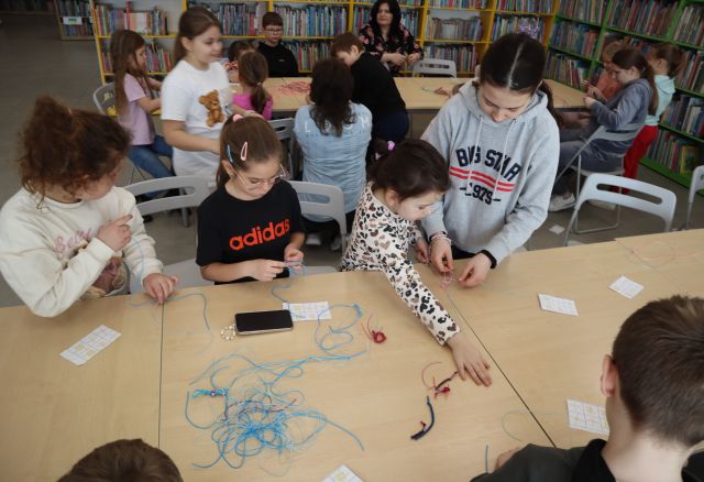 Na zdjęciu znajdują się dzieci przy stole, które robią breloczki z żyłki, jedna dziewczynka wyciąga rękę aby sięgnąć coś z końca stołu. W tle znajdują się złączone stoliki przy których uczestnicy również robią breloczki, a za nimi znajdują się półki z książkami.