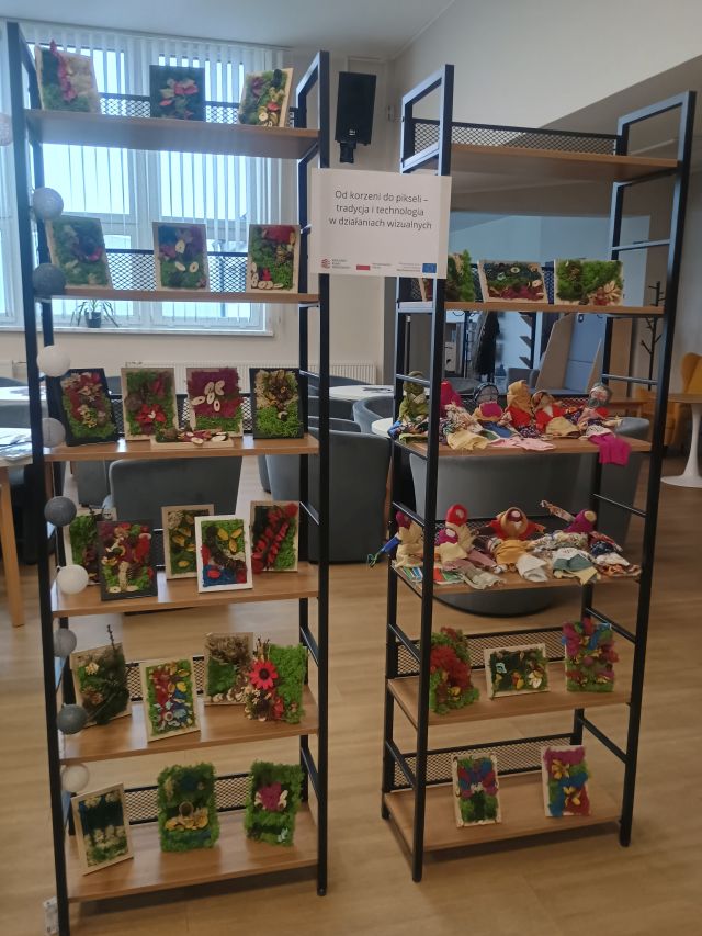 metalowe regały z drewnianymi półkami, na półkach obrazki z mchu i lalki motanki oraz nazwą projektu