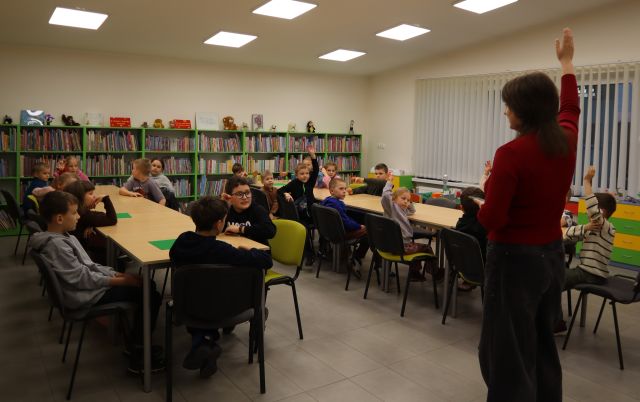 Na zdjęciu znajduje się kobieta z podniesioną ręką, które stoi pomiędzy złożonymi stołami w dwóch rzędach, przy których siedzą dzieci i część z nich również podnosi rękę do góry. W tle znajdują się półki z książkami.