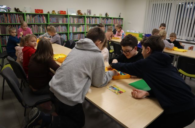 Na zdjęciu znajdują się dzieci przy złączonych stołach, w centrum znajduje się trójka chłopców, którzy układają LEGO. W tle znajduje się jeszcze jeden rząd ze złączonych stołów, przy którym siedzą dzieci. W tle znajdują się półki z książkami.