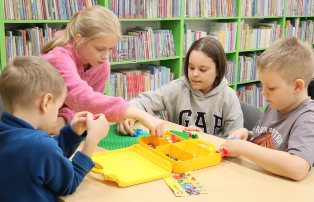 Na zdjęciu znajdują się dwie dziewczynki i dwóch chłopców, którzy wspólnie układają konstrukcje z LEGO, jedna dziewczynka nachyla się aby sięgnąć klocek z pudełka. W tle znajdują się półki z książkami.