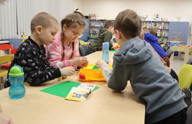 Na zdjęciu znajduje się trójka dzieci stojąca przy złączonych stołach i wspólnie układają LEGO, w tle znajdują się inni uczestnicy przy stole oraz półki z grami.