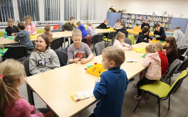 Na zdjęciu znajdują się dwa rzędy złączonych stołów, przy których siedzą dzieci i układają LEGO w grupach. W tle znajdują się okna.