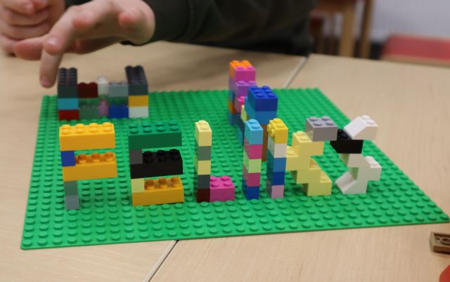 Na zdjęciu znajduje się zbudowany napis ,,FELIKS" z klocków LEGO na zielonej podkładce, na której znajdują się jeszcze dwie litery. Praca leży na stole.