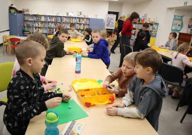 Na zdjęciu znajduje się rząd złączonych stołów przy którym siedzą dzieci i w małych grupach układają wspólnie LEGO, po prawej stronie znajduje się część drugiego stołu z resztą uczestników przy którym stoi kobieta. W tle znajdują się półki z książkami.