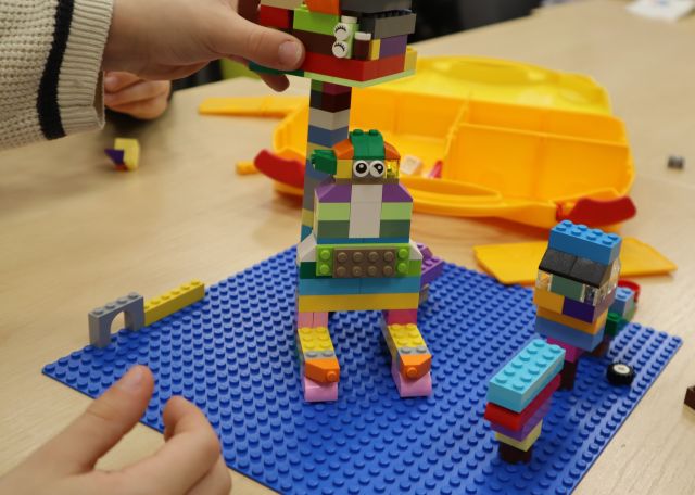 Na zdjęciu znajduje się robot zbudowany z klocków LEGO na podkładce, która leży na stole. W tle znajduje się walizeczka do przechowywania klocków.