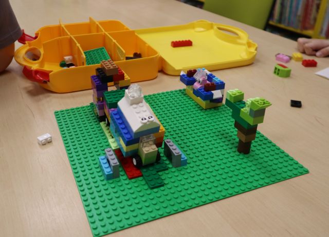 Na zdjęciu znajduje się budynek, drzewo kanapa zbudowane z klocków LEGO na zielonej podkładce, w tle znajduje się żółta walizeczka z klockami.