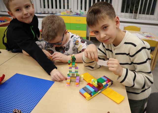 Na zdjęciu znajduje się trzech chłopców przy stole, dwóch uśmiecha się do zdjęcia wraz ze zbudowanym z klocków LEGO samolotem oraz robotem, które leżą przed nimi na stole. Chłopiec po środku skupiony jest na budowaniu. Na stole znajduje się również niebieska podkładka, a w tle znajdują się okna.