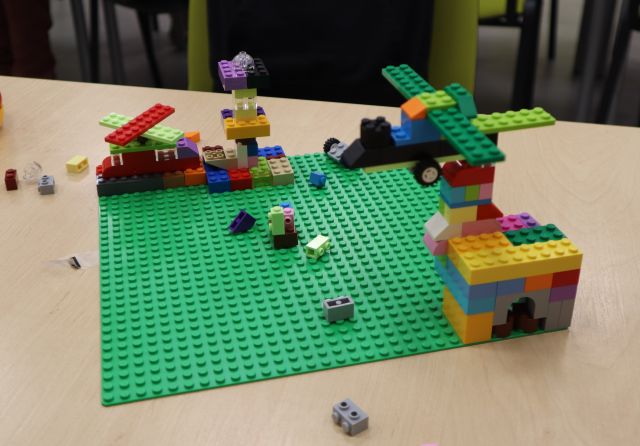Na zdjęciu znajduje się zielona plansza na stole, na której zbudowany jest mały samolot oraz część miasteczka z klocków LEGO.