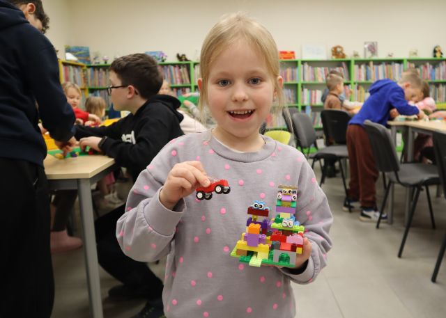 Na zdjęciu znajduje się uśmiechnięta dziewczynka, która stoi do zdjęcia wraz ze zbudowanymi przez siebie z klocków LEGO konstrukcjami. W tle znajdują się dwa rzędy połączonych stołów wraz z resztą uczestników oraz półki z książkami.