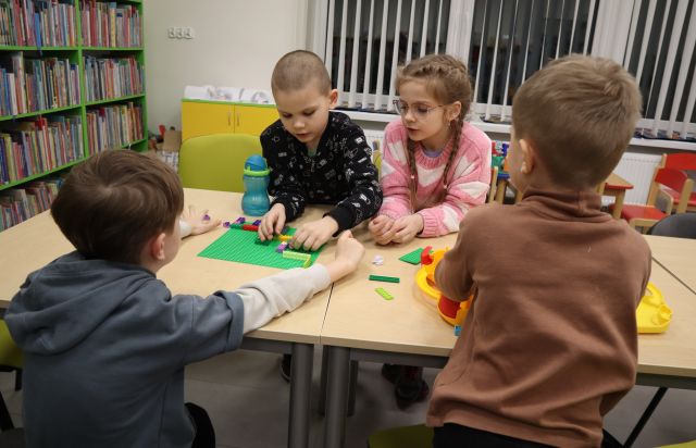 Na zdjęciu znajduje się czwórka dzieci siedząca na przeciwko siebie przy złączonych stołach i wspólnie układają labirynt z klocków LEGO znajdujących się na stole. W tle znajdują się okna oraz półki z książkami po lewej.