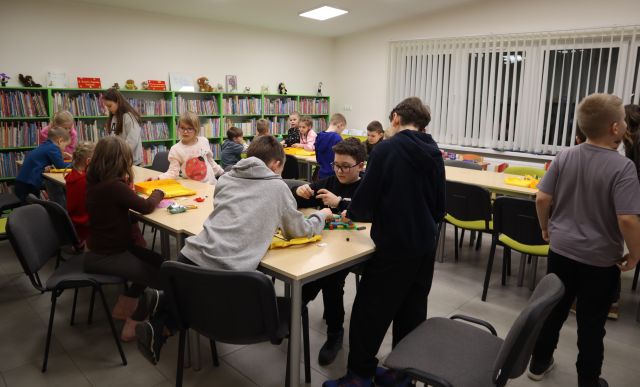 Na zdjęciu znajdują się dzieci przy złączonych w dwóch rzędach stołach i układają klocki LEGO, pomiędzy stołami, tyłem stoi chłopiec. W tle znajdują się półki z książkami oraz okna.