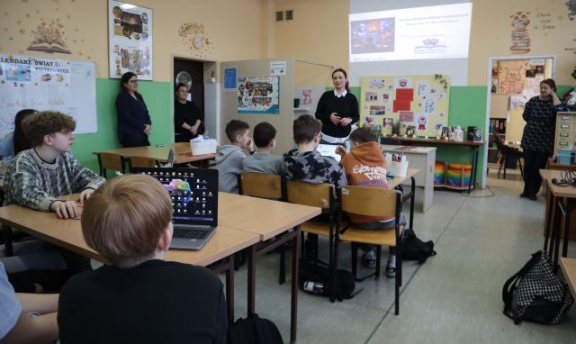 Na zdjęciu znajduje się młodzież przy stolikach przed nimi stoi kobieta prowadząca warsztaty, z boku stoi jeszcze jedna kobieta i opiera się o szafkę. Dwie kobiety stoją po lewej stronie i opierają się o ściane. Na stolikach uczniów leżą laptopy, a warsztaty odbywają się w szkolnej bibliotece.