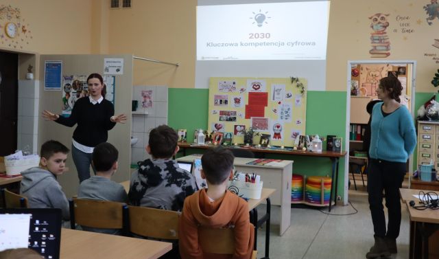 Na zdjęciu znajduje się czwórka uczniów przy stole, przed nimi stoją dwie kobiety, a na ścianie wyświetlana jest prezentacja. Warsztaty odbywają się w szkolnej bibliotece.