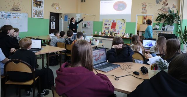Na zdjęciu znajduje się młodzież przy stolikach, na których leżą laptopy. Przed nimi stoją dwie kobiety i jedna ich wskazuje ręką w stronę wyświetlanej prezentacji. Warsztaty odbywają się w bibliotece szkolnej.