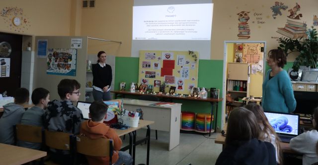 Na zdjęciu znajduje się czórka chłopców przy jednym stoliku i dwie dziewczyny przy stoliku po prawej. Przed nimi stoją dwie kobiety prowadzące zajęcia za nimi na ścianie wyświetlana jest prezentacja. Warsztaty odbywają się w szkolnej bibliotece.