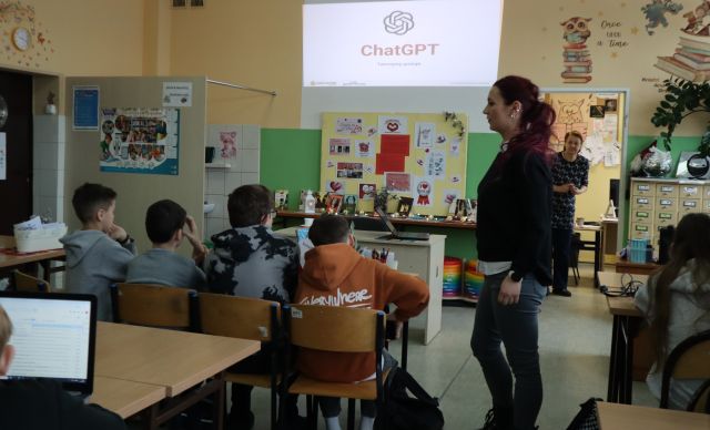 Na zdjęciu znajduje się młodzież przy stolikach, na której leżą laptopy. Pomiędzy stolikami stoi kobieta prowadząca zajęcia. Na ścianie wyświetlana jest prezentacja przedstawiająca ChatGPT. Zajęcia odbywają się w szkolnej bibliotece.