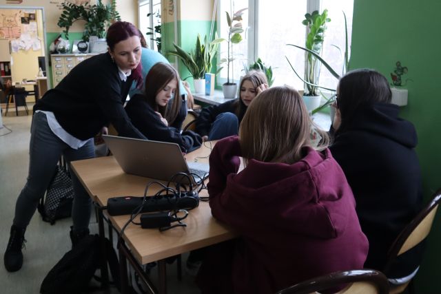 Na zdjęciu znajdują się cztery dziewczyny siedzące przy stole na przeciwko siebie. Na środku stołu leży laptop, do którego nachyla się kobieta prowadząca zajęcia. W tle znajdują się okna.