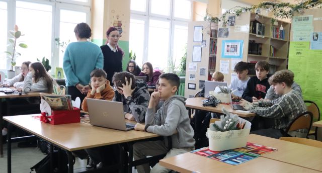Na zdjęciu znajdują się uczniowie podstawówki przy stolikach, na których znajdują się laptopy. Między stolikami stoją dwie kobiety. Warsztaty odbywają się w sali komputerowej.
