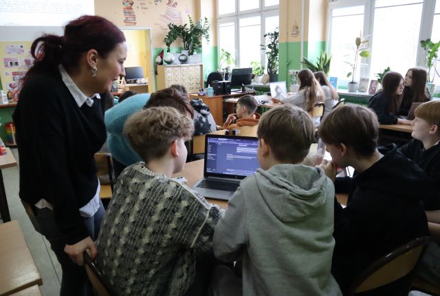 Na zdjęciu znajduje się czwórka chłopców przy stoliku, po lewej stronie nachyla się kobieta . Przed nimi na stole leży laptop, a w tle znajdują się stoliki z resztą uczestników oraz okna.