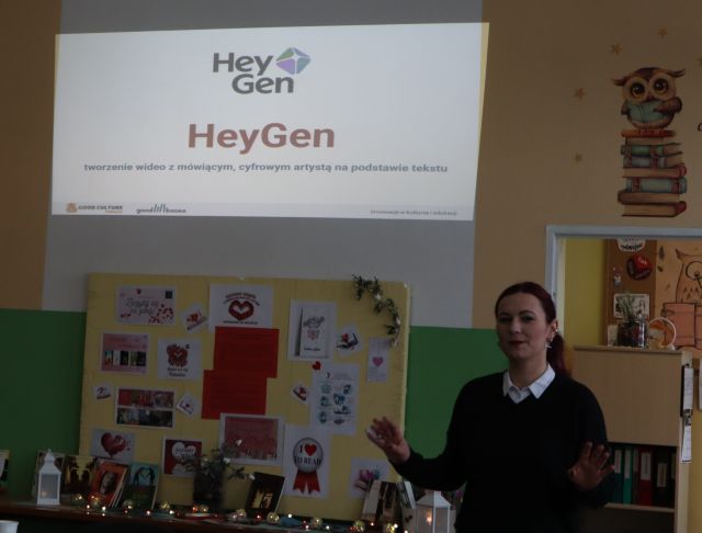 Na zdjęciu po prawej stronie znajduje się kobieta od pasa w górę, obok niej na ścianie wyświetlana jest prezentacja o aplikacji HeyGen. Warsztaty odbywają się w szkolnej bibliotece.