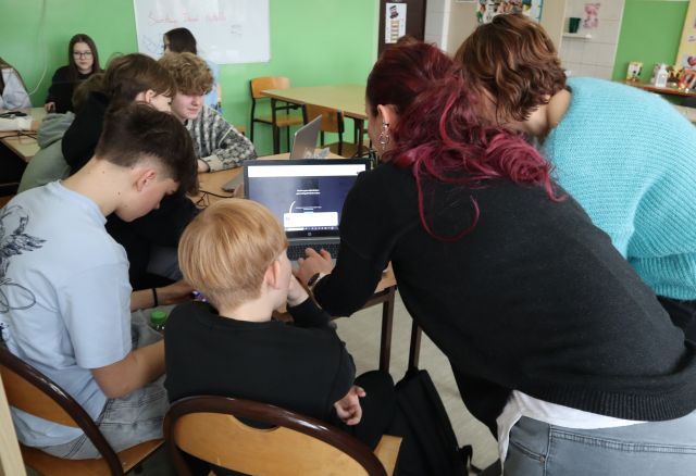 Na zdjęciu znajduję się grupka chłopaków przy stoliku z laptopem oraz dwie kobiety prowadzące zajęcia, które się nachylają nad laptopem. W tle znajdują się stoliki z uczestnikami warsztatów.