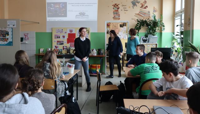 Na zdjęciu znajdują się uczniowie podstawówki przy stolikach, a przed nimi stoją trzy kobiety. W tle na ścianie wyświetlana jest prezentacja. Warsztaty odbywają się w sali komputerowej.