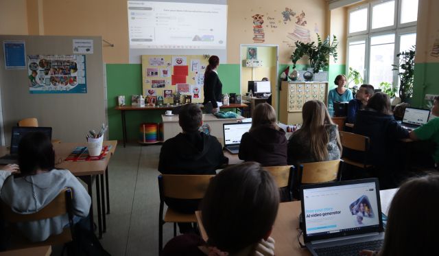 Na zdjęciu znajduję się młodzież przy stolikach z laptopami, przed nimi znajduje się kobieta prowadząca zajęcia. Warsztaty odbywają się w szkolnej bibliotece.