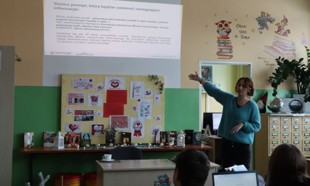 Na zdjęciu znajduje się kobieta wskazująca na prezentację. Warsztaty odbywają się szkolnej bibliotece.