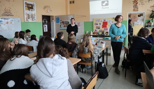 Na zdjęciu znajdują się uczniowie przy stolikach z laptopami. Przed nimi stoją dwie kobiety a za nimi wyświetlana jest prezentacja. Warsztaty odbywają się w szkolnej bibliotece.