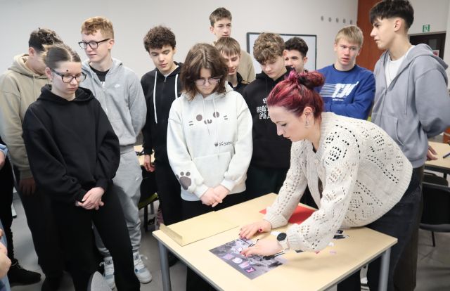 Na zdjęciu znajduje się stolik z kartkami i zdjęciami przy którym stoi młodzież, a po prawej stronie nachyla się kobieta i ustawia zdjęcia. Warsztaty odbywają się w sali komputerowej.