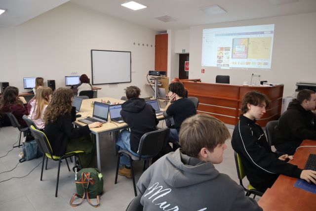 Na zdjęciu znajdują się uczniowie przy złączonych w prostokąt stołach oraz przy komputerach. Na ścianie wyświetlana jest prezentacja. Warsztaty odbywają się w sali komputerowej.