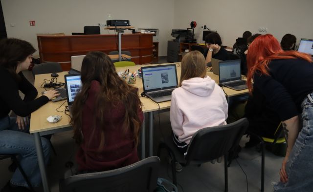 Na zdjęciu znajdują się stoły złączone w prostokąt przy których siedzi młodzież wraz z laptopami, na których wykonują kolaż, nad jedną z osób po lewej stronie znajduje się nachylająca się kobieta prowadząca zajęcia. Zajęcia odbywają się w sali komputerowej.