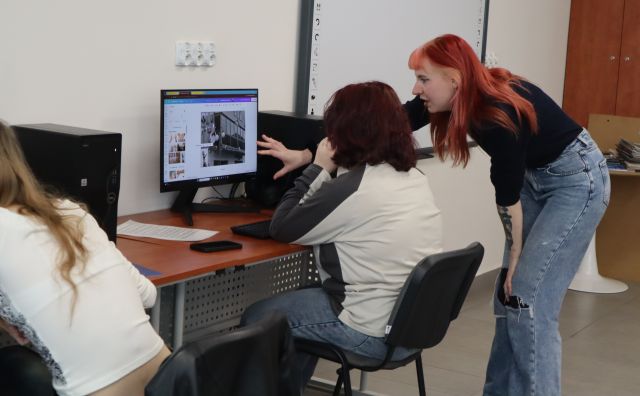 Na zdjęciu znajduje się dziewczyna siedząca przy komputerze, a koło niech nachyla się kobieta prowadząca zajęcia i wskazuje ręką na ekran, na którym znajduje się kolaż stworzony przez nastolatkę. Po lewej stronie zdjęcia znajduje się siedząca dziewczyna. Warsztaty odbywają się w sali komputerowej.