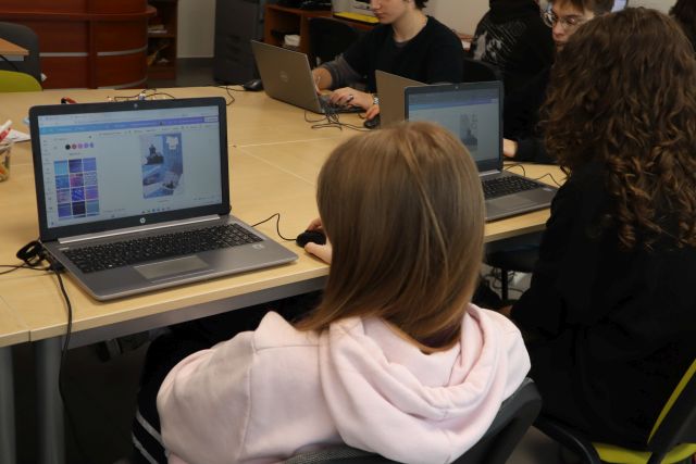Na zdjęciu w centrum znajduje się dziewczyna siedząca przed laptopem, który leży na stole i kończy swój kolaż dotyczący miasta. Obok niej siedzi inna uczestniczka zajęć. Zajęcia odbywają się w sali komputerowej, a zdjęcie zostało wykonane od tyłu.