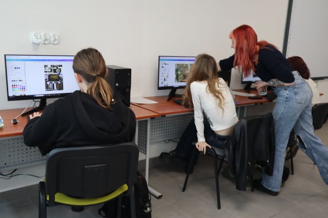 Na zdjęciu znajdują się trzy osoby siedząca przy komputerach oraz kobieta nachylajaca sie przy drugiej osobie i pokazuje coś na ekranie. Warsztaty odbywają się w sali komputerowej.