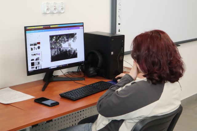 Na zdjęciu znajduje się dziewczyna przy komputerze, która kończy swój kolaż, na którym dominuje kolor czarny. Warsztaty odbywaja się w sali komputerowej.