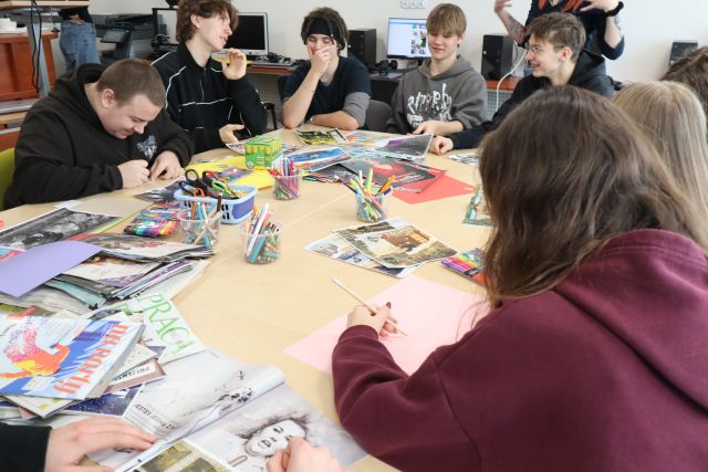 Na zdjęciu znajduje się młodzież przy złączonych stolikach, przed nimi znajdują się kolorowe kartki, gazety, nożyczki oraz kleje. Warsztaty odbywają się w sali komputerowej.