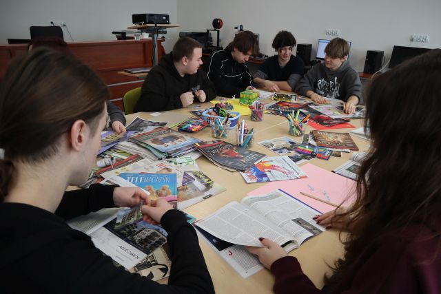 Na zdjęciu znajduja się uczniowie przy złączonych stołach, na których znajdują sie kartki i gazety, z których wykonują kolaże. Warsztaty odbywają się w sali komputerowej.