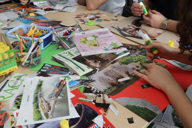 Na zdjęciu znajdują się gazety, kolorowe kartki, nożyczki, kleje i kredki leżące na stole. Przy stole znajdują się ręce uczestników warsztatów, którzy kleją i tworzą swoje własne kolaże.
