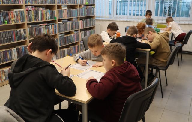 Na zdjęciu znajdują się dzieci przy trzech stołach i mają przed sobą kartkę. W tle znajdują się półki z książkami.