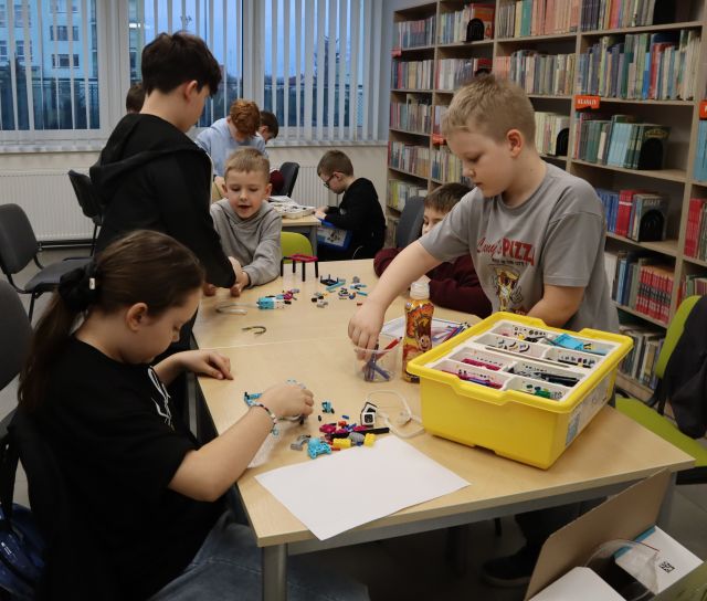 Na zdjęciu znajduje się piątka dzieci przy złączonych stolikach i wspólnie układają LEGO. W tle znajduje się reszta uczestników zajęć oraz półki z książkami po prawej stronie.