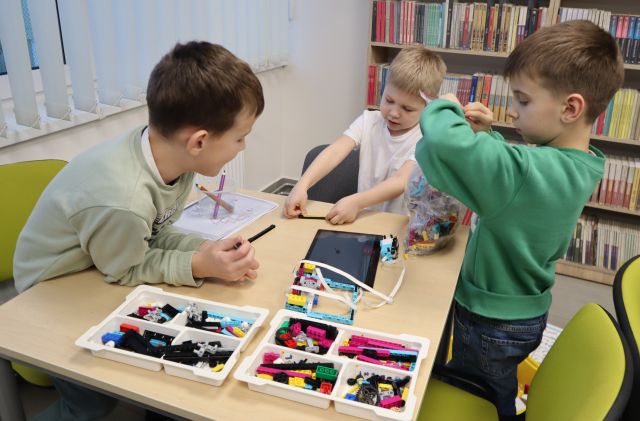 Na zdjęciu znajduje się trójka chłopców przy stole, jeden z nich stoi. Na stoliku leży LEGO, które wspólnie budują oraz tablet, na którym wyświetlana jest instrukcja. W tle znajdują się półki z książkami i okna po lewej stronie.